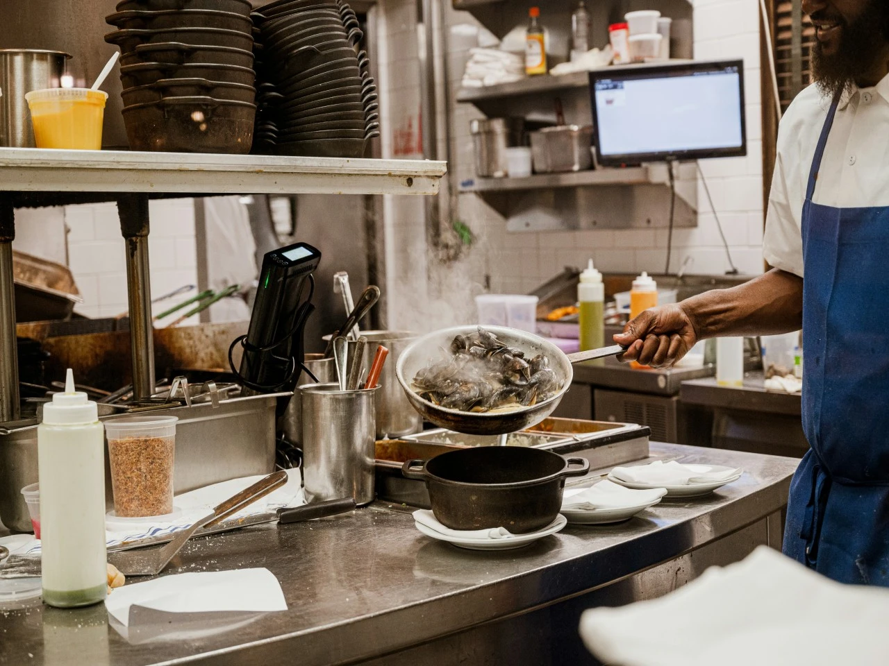A cook plating a dish at the kitchen pass beside a display screen